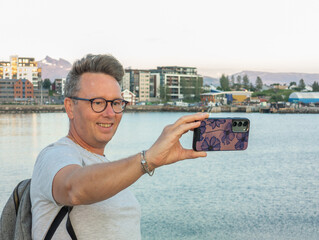Caucasian tourist taking a selfie with his smartphone during evening in Bodø, Norway. Yachts in the harbor of Bodo in the background. Norwegian city, solo traveller taking photo with cellphone.