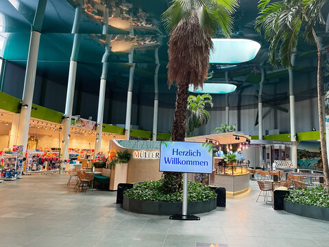 Herzlich Willkommen at a modern German Therme Erding mall atrium with palm trees, shopping stalls, and cafe