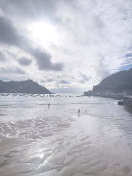 Playa con arena h&uacute;meda y reflejos de luz frente a bah&iacute;a con barcas y colinas, bajo cielo parcialmente nublado.