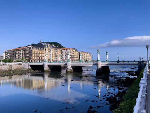 Puente con pilares ornamentados sobre estuario en calma, flanqueado por edificios cl&aacute;sicos y barandilla blanca bajo cielo azul.