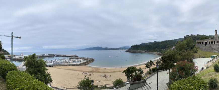 Bah&iacute;a tranquila con playa amplia, zona de puerto con barcos y gr&uacute;a, y colina verde con estructura de piedra dominando la costa bajo cielo nublado.
