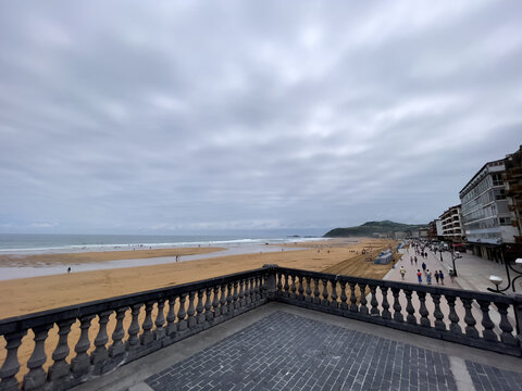 Terraza de piedra con balaustrada frente a playa amplia, gente paseando junto a edificios y colina verde al fondo bajo cielo nublado.