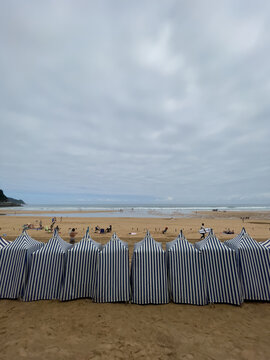 Cabinas rayadas alineadas sobre la arena, gente descansando frente al mar y cielo nublado sobre olas suaves.