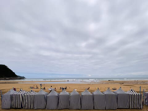 Cabinas rayadas alineadas sobre la arena, gente descansando frente al mar y colina oscura al fondo bajo cielo nublado.