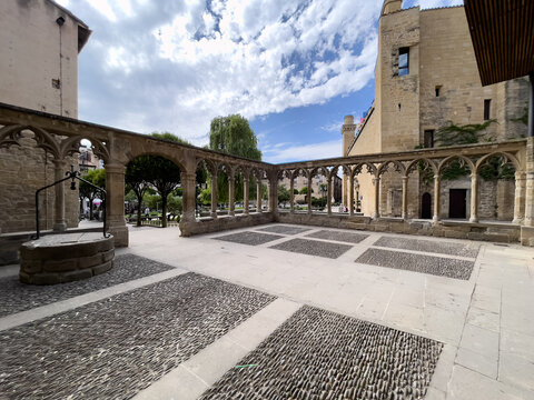 Edificio hist&oacute;rico de piedra con torre campanario y torrecillas de tejado c&oacute;nico, rodeado de patio empedrado y &aacute;rboles bajo cielo azul con nubes dispersas.