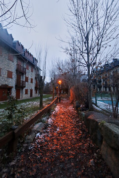 Sendero con hojas y nieve, iluminado por una farola c&aacute;lida entre &aacute;rboles desnudos y casas de piedra con balcones.