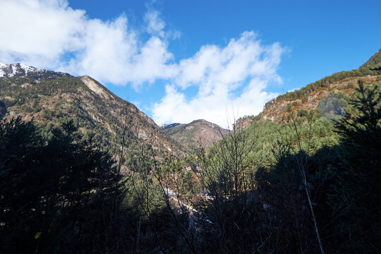 Laderas verdes y boscosas ascendiendo hacia picos rocosos, bajo un cielo azul con nubes blancas.