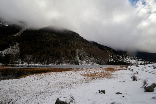 Campo nevado junto a una l&aacute;mina de agua tranquila, con laderas boscosas cubiertas de niebla bajo un cielo gris con claros.
