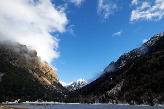 Valle de monta&ntilde;a con cumbres nevadas y laderas boscosas entre nubes bajas, bajo un cielo azul que aporta claridad al paisaje invernal.