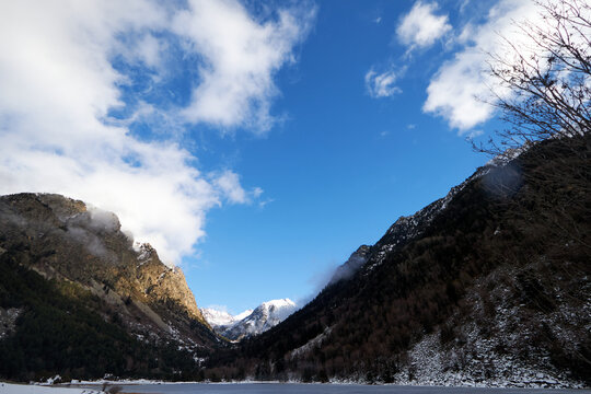 Valle de monta&ntilde;a con laderas nevadas y un pico iluminado al fondo, bajo un cielo azul con nubes dispersas que realzan el contraste del paisaje invernal.