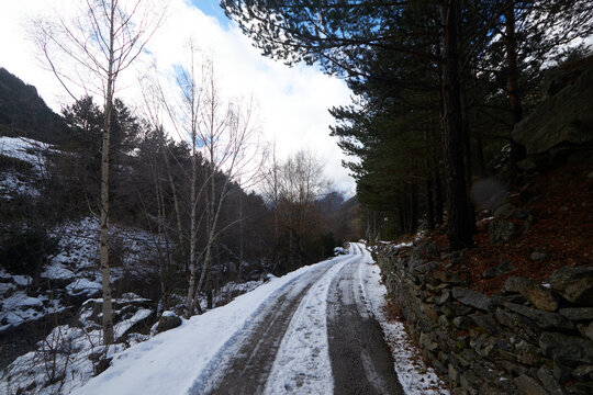 Sendero estrecho cubierto de nieve que avanza entre un muro de piedra y un bosque de con&iacute;feras, con &aacute;rboles desnudos junto a un peque&ntilde;o arroyo y luz suave filtrada por nubes que aporta una atm&oacute;sfera t