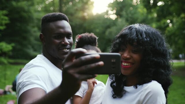 african american family having fun in the park pose to take a selfie with mobile phone waving hand slow motion close up child girl daughter couple summer nature picnic mother parents portrait