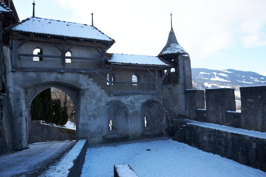 Castillo de piedra con arco y torre nevada, pasarela de madera y monta&ntilde;as blancas bajo cielo claro.