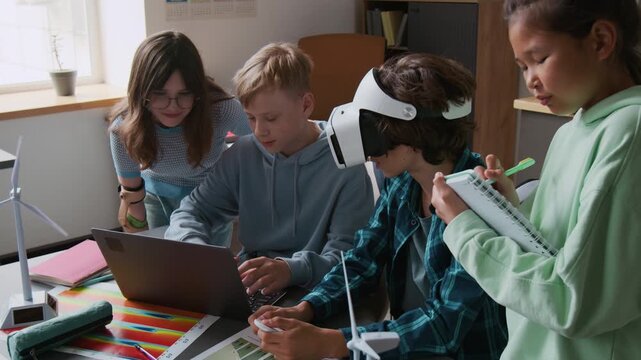 High angle shot of group of children using laptop and VR glasses while working on renewable energy project together in school classroom