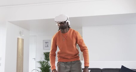 Donning white VR headset, African American man raising and exploring virtual space in living room - Powered by Adobe