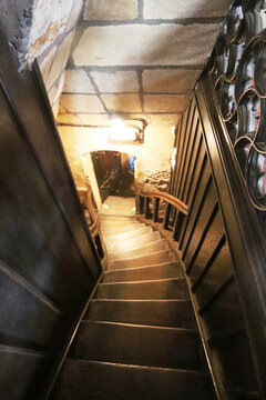 Escalera estrecha y empinada de madera oscura que desciende entre muros de piedra, con barandilla de hierro forjado y una luz c&aacute;lida al fondo que ilumina suavemente el descenso.