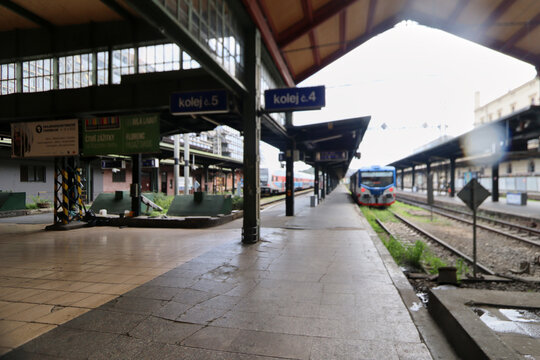And&eacute;n de estaci&oacute;n con varias v&iacute;as, marquesinas met&aacute;licas, se&ntilde;alizaci&oacute;n de andenes y un tren azul, blanco y rojo detenido bajo un cielo gris.