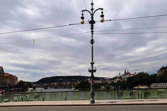 Puente urbano con farola ornamental de doble esfera en primer plano, un r&iacute;o fluyendo entre edificios y zonas verdes, y colinas con una torre al fondo bajo un cielo gris.