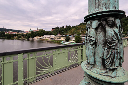 Detalle de un puente con columna met&aacute;lica verde con relieves figurativos y barandilla ornamental, frente a un r&iacute;o flanqueado por edificios y laderas verdes bajo un cielo nublado.