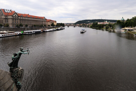 R&iacute;o amplio con barcos navegando, un edificio hist&oacute;rico de tejado rojo y una estatua de bronce en primer plano, con un puente al fondo bajo un cielo gris.