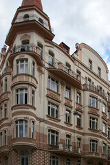 Obraz premium Edificio histórico de tonos crema y rojizos con molduras decorativas, balcones de hierro forjado y torre esquinera de tejado puntiagudo, destacado sobre un cielo parcialmente nublado.