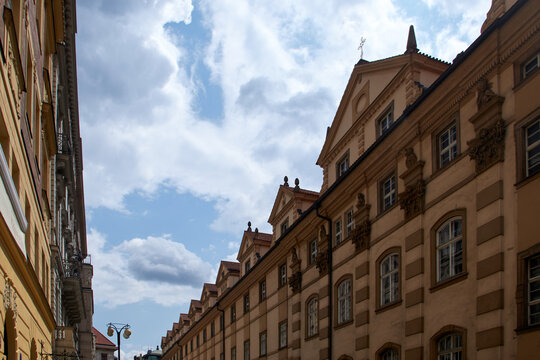 Calle estrecha flanqueada por edificios hist&oacute;ricos en tonos beige y marr&oacute;n claro, con molduras ornamentadas y tejados inclinados, bajo un cielo parcialmente nublado.
