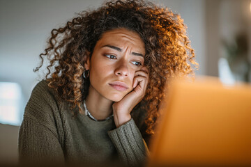 Young woman with curly hair looks stressed while working on a laptop in a cozy indoor setting during evening hours