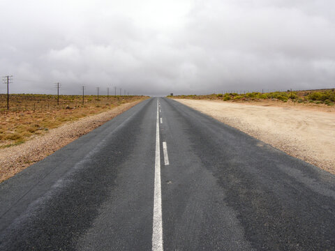 Carretera asfaltada que se pierde en el horizonte entre terrenos &aacute;ridos, con postes telef&oacute;nicos a un lado y un cielo gris cargado de nubes.