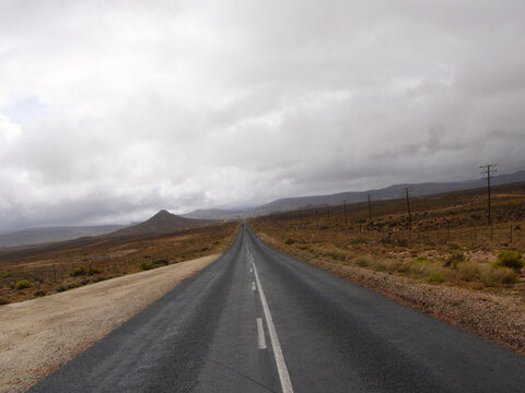 Carretera recta que atraviesa un paisaje semi&aacute;rido con llanuras secas, vegetaci&oacute;n dispersa y l&iacute;neas el&eacute;ctricas paralelas, rumbo a colinas lejanas bajo un cielo nublado.