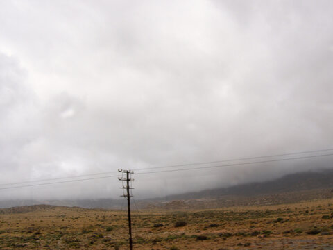 Terreno &aacute;rido con vegetaci&oacute;n dispersa y un poste el&eacute;ctrico en primer plano, frente a colinas parcialmente cubiertas por nubes grises que anuncian mal tiempo.