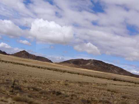 Paisaje &aacute;rido con matorrales dispersos y colinas redondeadas de roca al fondo, bajo un cielo parcialmente nublado.