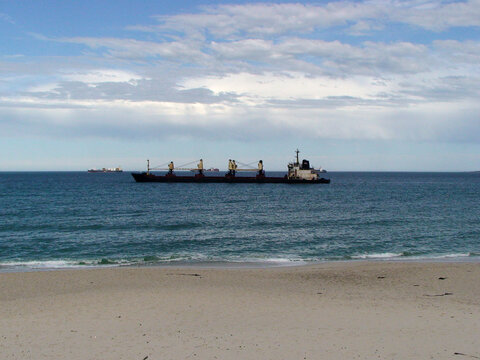 Buque de carga navegando cerca de una playa de arena, con gr&uacute;as en cubierta y mar en calma bajo un cielo parcialmente nublado.