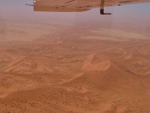 Vista a&eacute;rea de un desierto de dunas rojizas con relieves ondulados, captado desde un avi&oacute;n cuya ala aparece en la parte superior, bajo un cielo claro.