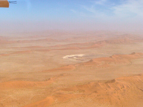 Vista a&eacute;rea de un desierto rojizo con un gran c&iacute;rculo claro que parece una cuenca seca o salar, rodeado de terreno arenoso y rocoso bajo un cielo azul tenue.
