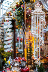 A close-up of a hanging wind chime made of shells and beads at a market stall. Christmas lights and other decorations are in the blurred background.