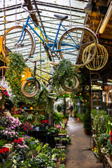 A narrow aisle inside a covered market. Potted plants and flowers are on both sides, with a hanging bicycle and a small children's bike decorated with greenery.