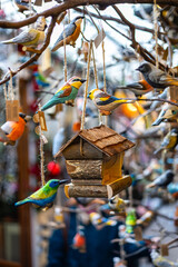 A wooden birdhouse with a thatched roof, surrounded by small, colorful, hand-painted wooden birds. The decorations are hanging from branches at a market stall.