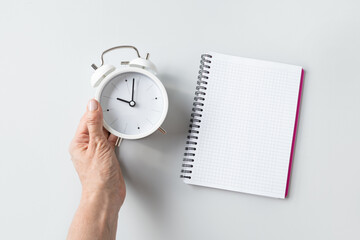 Alarm clock in woman hand and empty sheet blank on gray background. Daily routine. View from above....