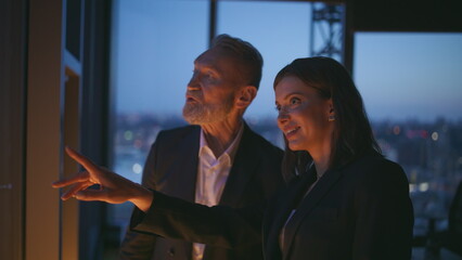 Two late workers standing by panoramic window business center closeup smiling 