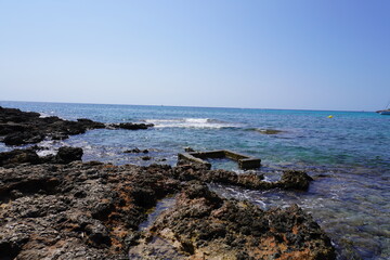 Felsige Küste mit türkisfarbenem Meer, Wellen und Überresten eines alten Betonfundaments im Wasser