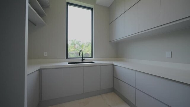 Modern kitchen or utility room with light gray handle less cabinets and white L shaped countertop. Black faucet and sink sit below tall window that brings in natural light