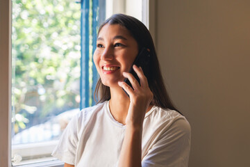 Young Asian woman happily talking on a smartphone while standing near a window with natural light