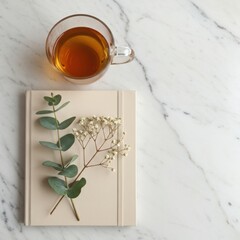 A clear glass mug of amber tea rests beside a closed beige notebook adorned with eucalyptus and baby s breath isolated on white background