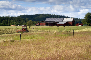 Fototapeta premium Red Tractor Harvesting Hay in Rural Scandinavian Landscape with Traditional Farm Buildings, Dollar Panels, and Countryside Backdrop Near Håverud, Sweden