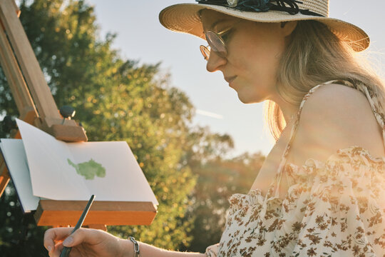 Young woman painter in sunlit park, creating art on easel.  Golden hour light enhances scene. Perfect for articles about art, nature, creativity, and summer.