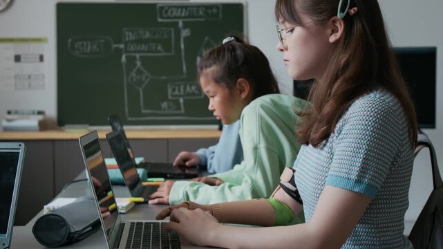 Tilt-up shot of teenage girl with prosthetic arm and her Asian classmate sitting together at desk and working on their laptops during IT class in school