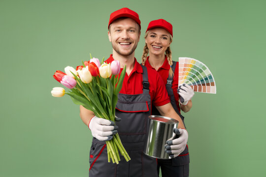 Portrait of painters with paint bucket, flowers bouquet and color swatches. Young adult couple holding paint can, color palette fan on green background. Painting service, paint shop, spring renovation