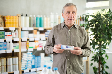 Focused older man in olive button-up shirt standing in pharmacy, examining box of ointment while choosing safe medicine for joint or back pain before making purchase..