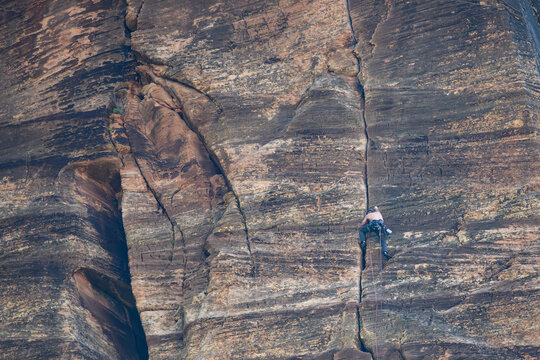 Recreational rock climber, shirtless man with safety helmet, harness, and rope, high up on a steep vertical cliff rock wall, Zion National Park, Utah
