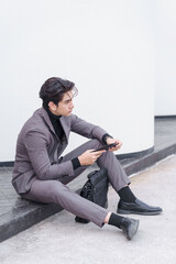 Young businessman sitting outdoors using smartphone with briefcase
